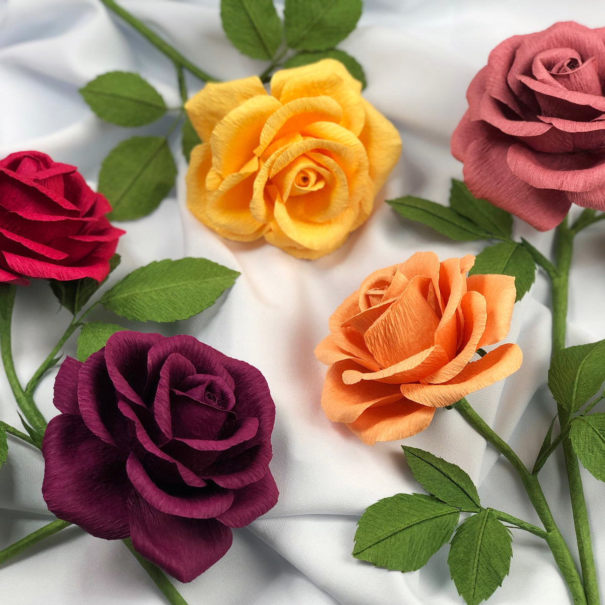 Red, Yellow, Pink, Orange and Dark Purple paper roses lying across white draped fabric