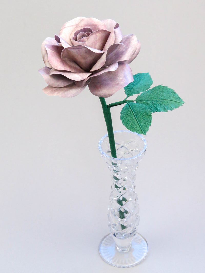 Grey tin paper rose with three forest green leaves standing in a narrow glass vase against a light grey backdrop