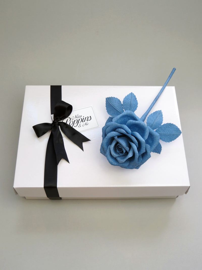 China blue crepe paper rose lying diagonally on top of a luxury white gift box that has a black satin ribbon tied in a bow with a Miss Poppins and Me gift tag attached