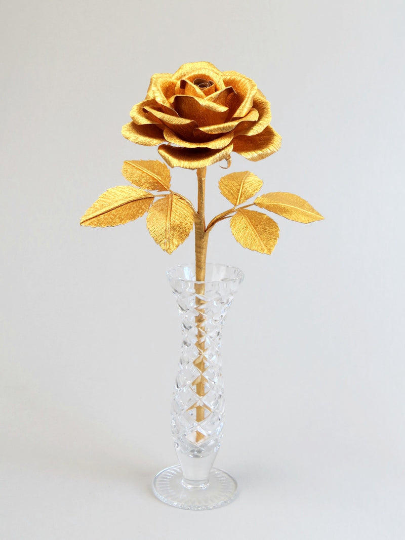 Gold crepe paper rose with six gold leaves standing in a narrow glass vase set against a light grey background