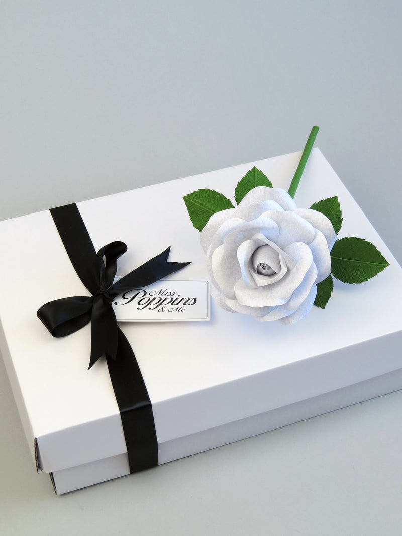 White lace printed paper rose lying diagonally on top of a luxury white gift box that has a black satin ribbon tied in a bow with a Miss Poppins and Me gift tag attached