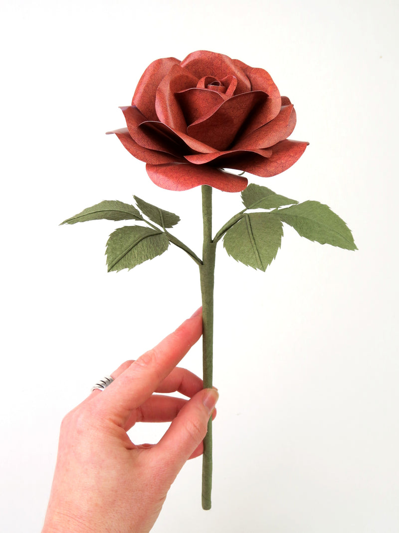 Pale white hand delicately holding the stem of a brown leather grain paper rose with six olive green leaves