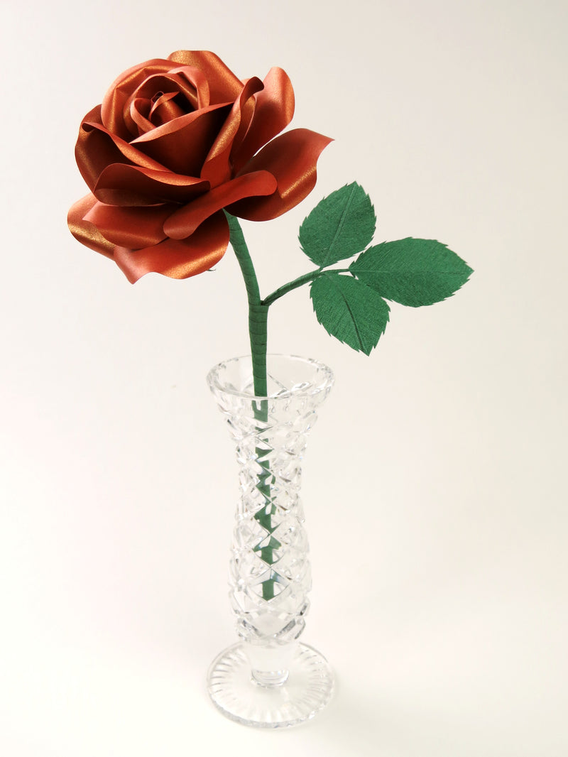 Copper paper rose with three leaves standing in a narrow glass vase against a light grey backdrop