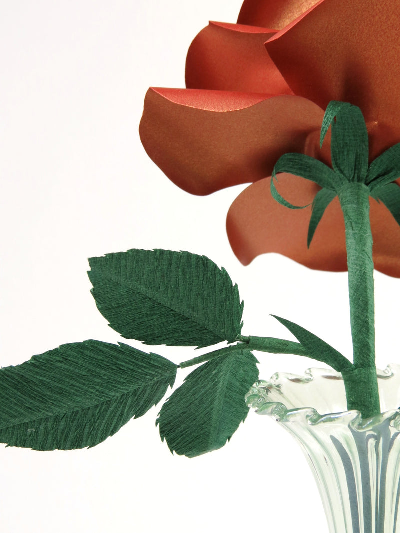 Detailed view of the back of a copper paper rose with a green curled calyx underneath the metallic petals, and the green stem, leaves and the tip of the glass vase just in view