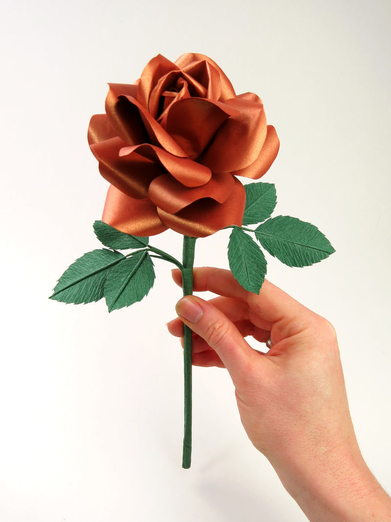 Pale white hand delicately holding the stem of a copper paper rose with six leaves