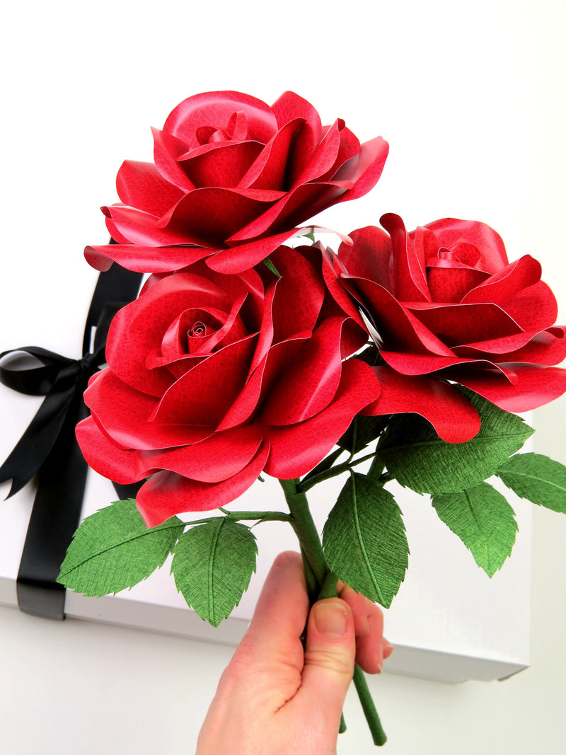 Pale white hand holding a bouquet of three red leather grain paper rose with ivy green leaves above a white gift box with a black satin ribbon bow
