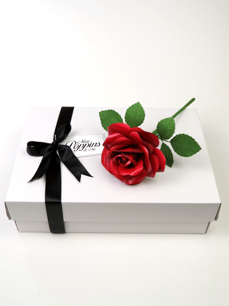 Red leather grain paper rose lying on top of a luxury white gift box that has a black satin ribbon tied in a bow with a Miss Poppins and Me gift tag attached