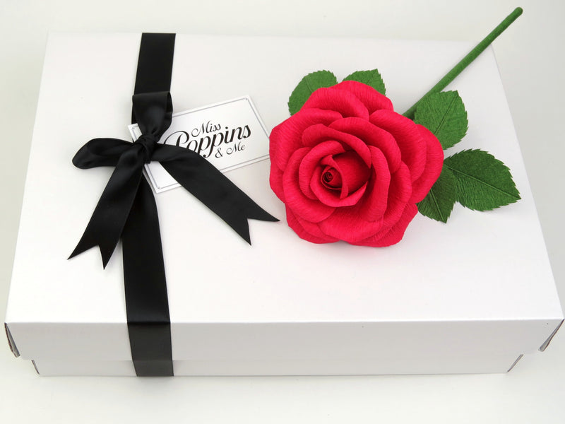 Red paper rose lying on top of a luxury white gift box that has a black satin ribbon tied in a bow with a Miss Poppins and Me gift tag attached