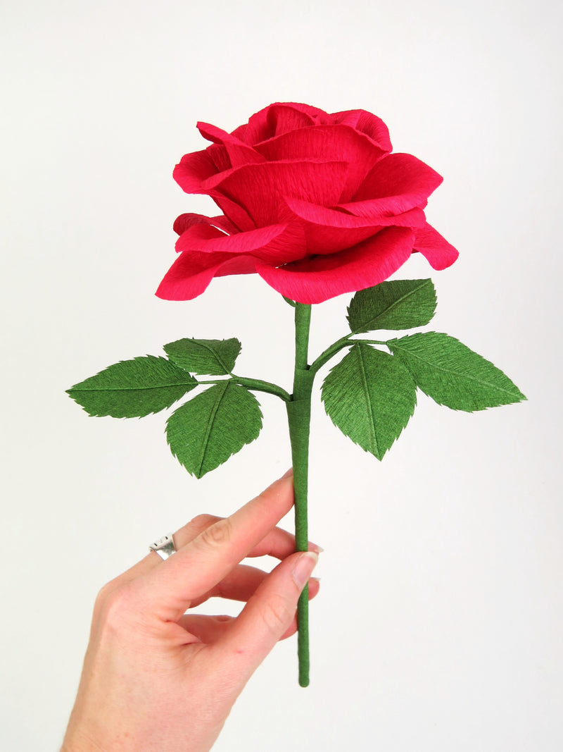 Pale white hand delicately holding the stem of a red paper rose with six leaves
