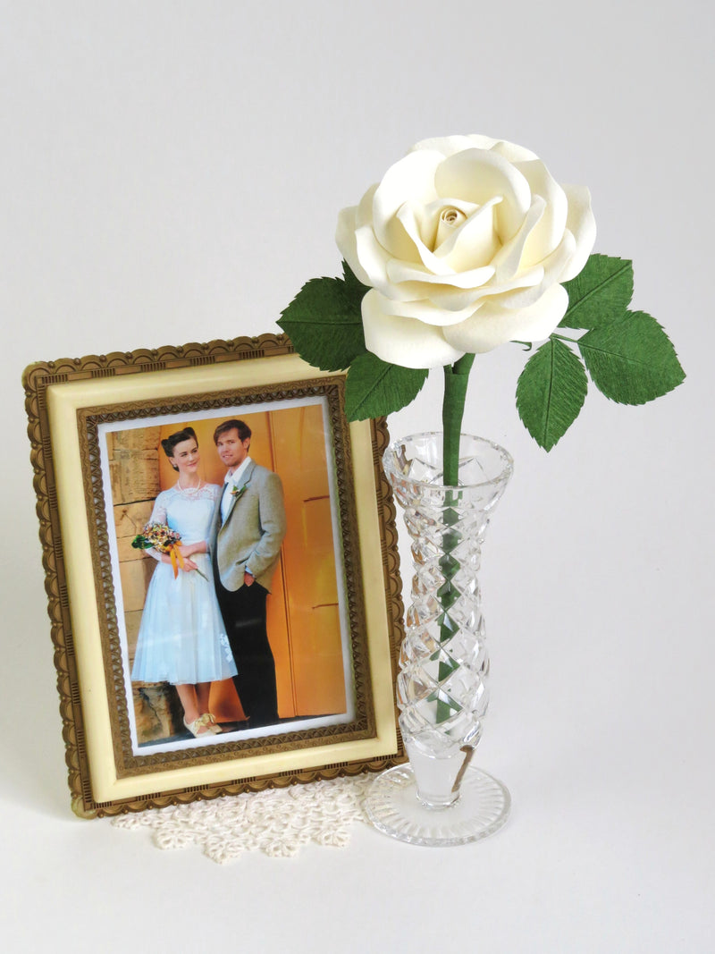White cotton paper rose with six green leaves standing in a slender glass vase with a cream and gold vintage framed wedding photo of a bride and groom