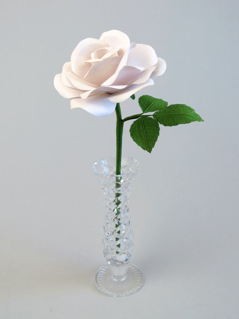 White linen grain paper rose with three leaves standing in a narrow glass vase against a light grey backdrop