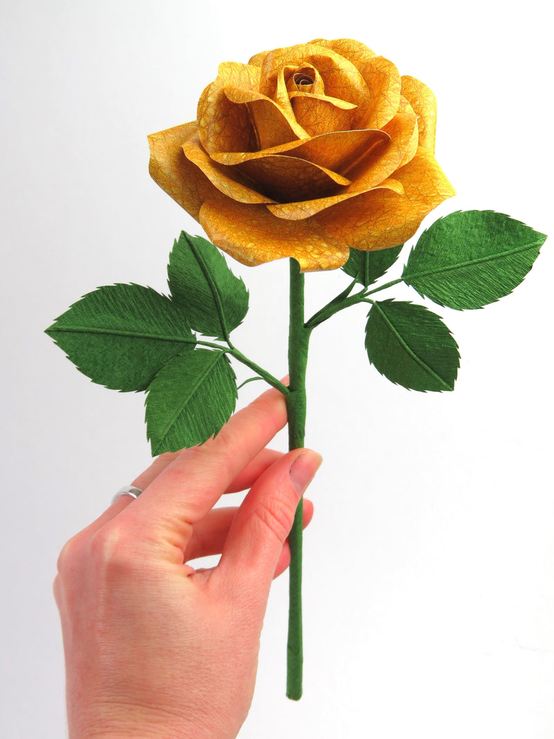 Pale white hand delicately holding the stem of a yellow leather grain paper rose with six leaves