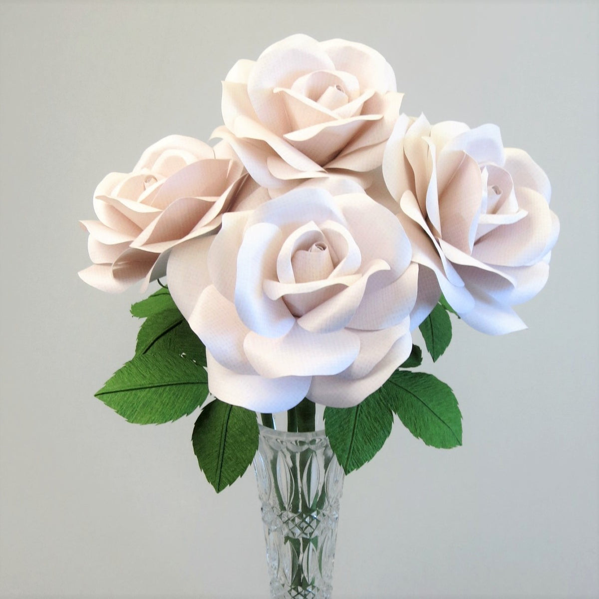 Bouquet of four white linen grain paper roses sitting tightly in a crystal vase with some ivy green leaves showing, set against a light grey background