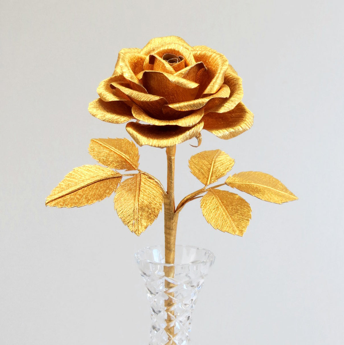 Glimmering gold rose standing upright in a slender crystal vase, with 6 gold leaves attached to the rose stem, standing against a grey background
