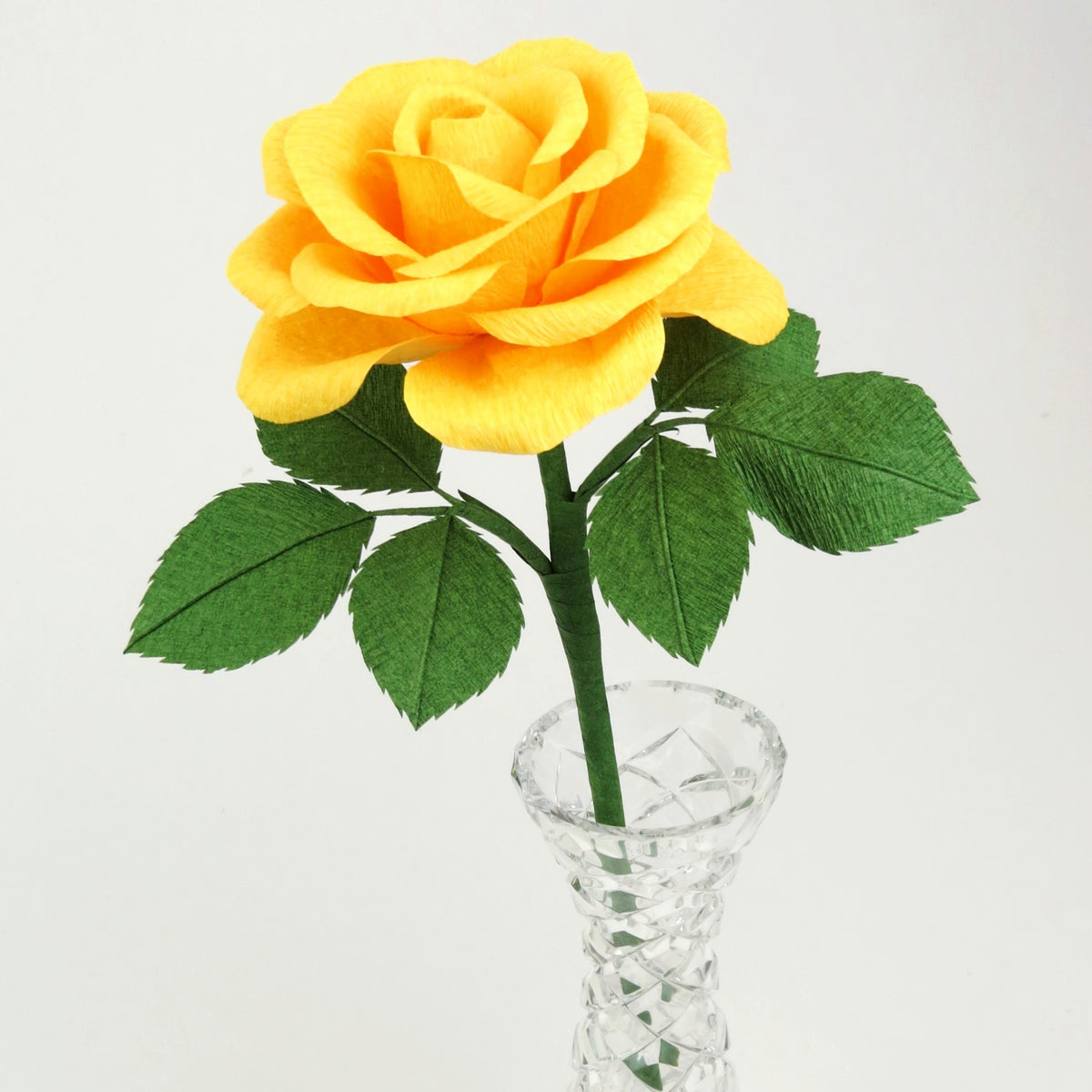 Yellow paper rose with six leaves standing in a narrow glass vase against a light grey background