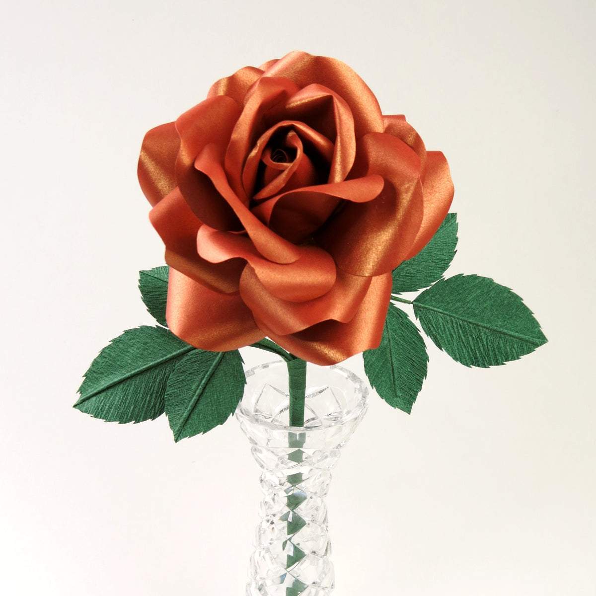 Copper paper rose with six green leaves standing in a narrow glass vase set against a light grey background