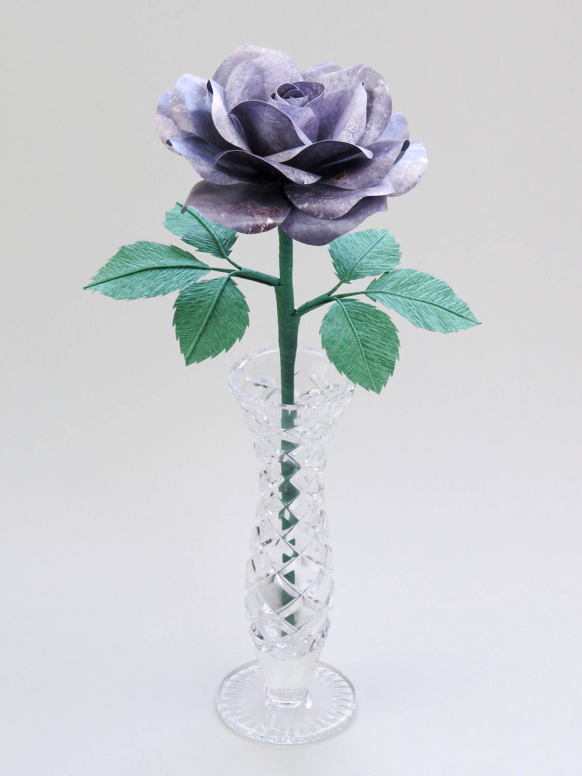 Grey iron paper rose with six forest green leaves standing in a narrow glass vase set against a light grey background