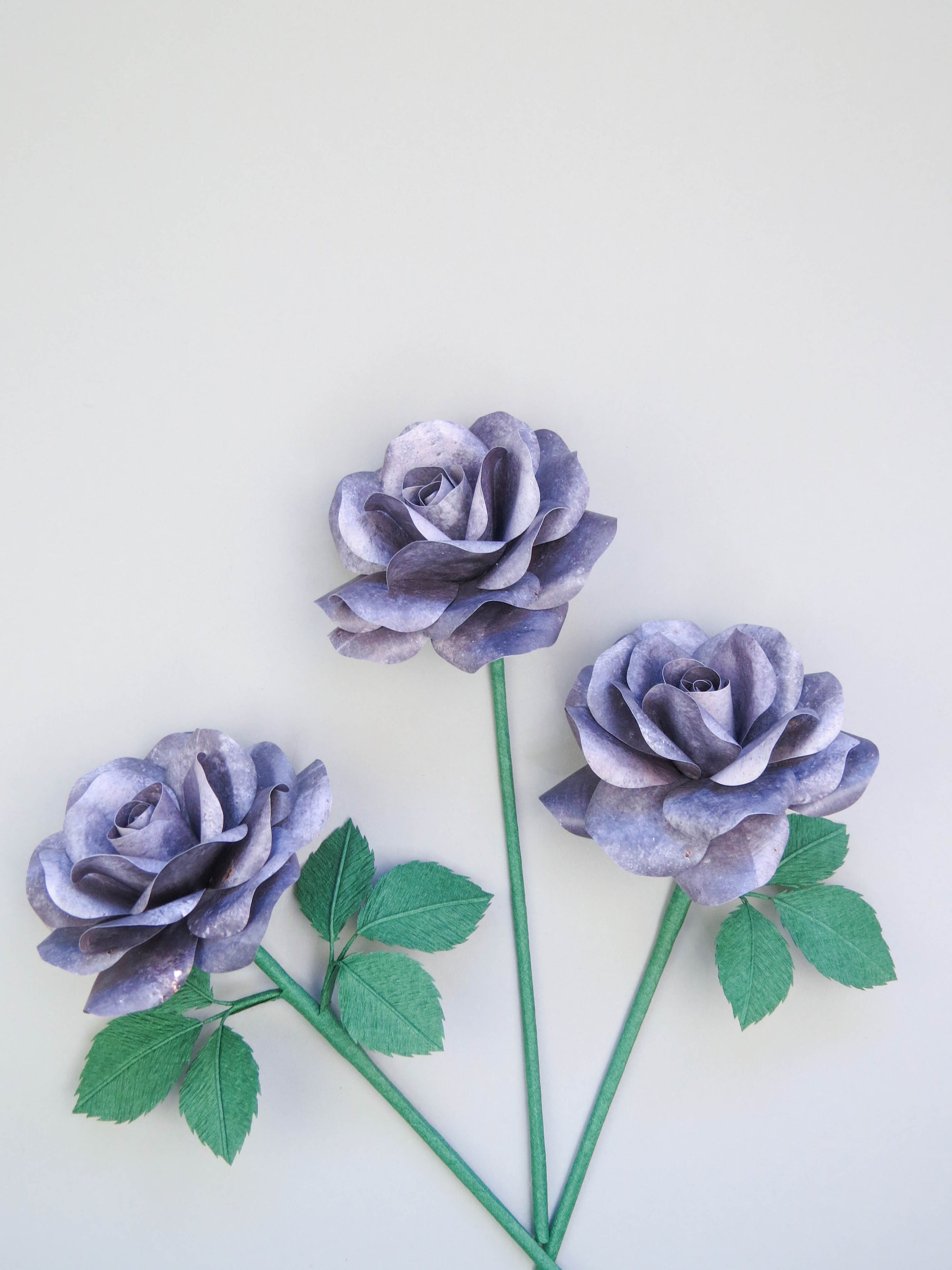 Three grey iron paper roses randomly lying next to each other on a light grey background. The left rose has six green leaves attached, the middle rose has no leaves and the right rose has three leaves.
