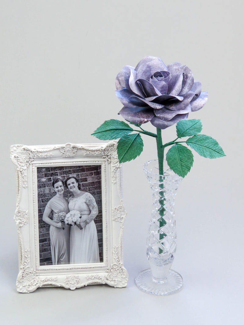 Grey iron paper rose with six forest green leaves standing in a slender glass vase with a white ornate framed black and white wedding photo of a happy bride and bridesmaid standing beside it