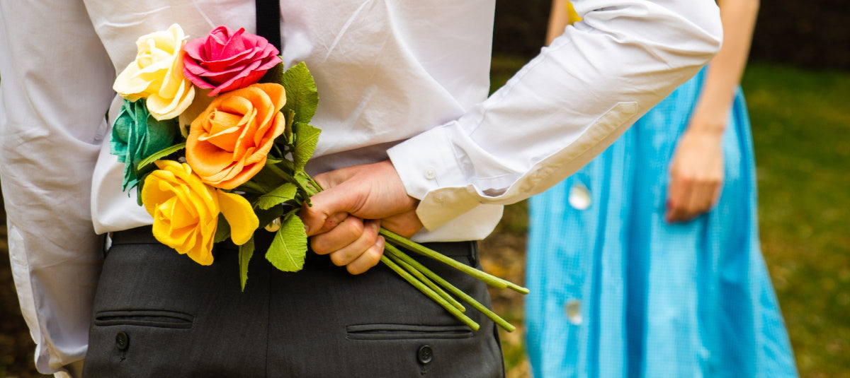 Bouquet of bright coloured paper roses hidden behind a man's back with a lady in a blue dress blurred and standing in front of him expectantly