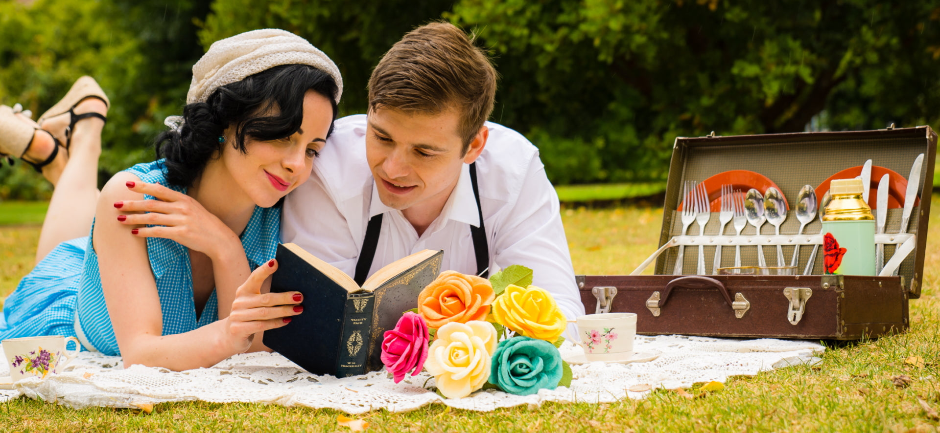 Vintage lady in sleeveless blue dress lying on a white crochet picnic blanket reading an old book with a gentleman lying next to her. There is a bright bouquet of paper flowers in front of them and a vintage picnic suitcase to the right of them. 