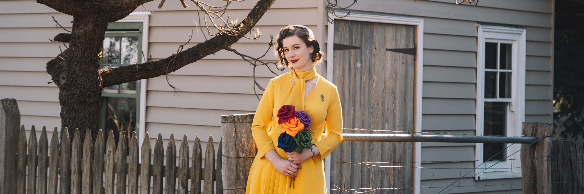 Miss Poppins wearing a bright yellow swing dress, holding a bright rainbow bouquet of paper roses against a rustic timber farms shed and gate