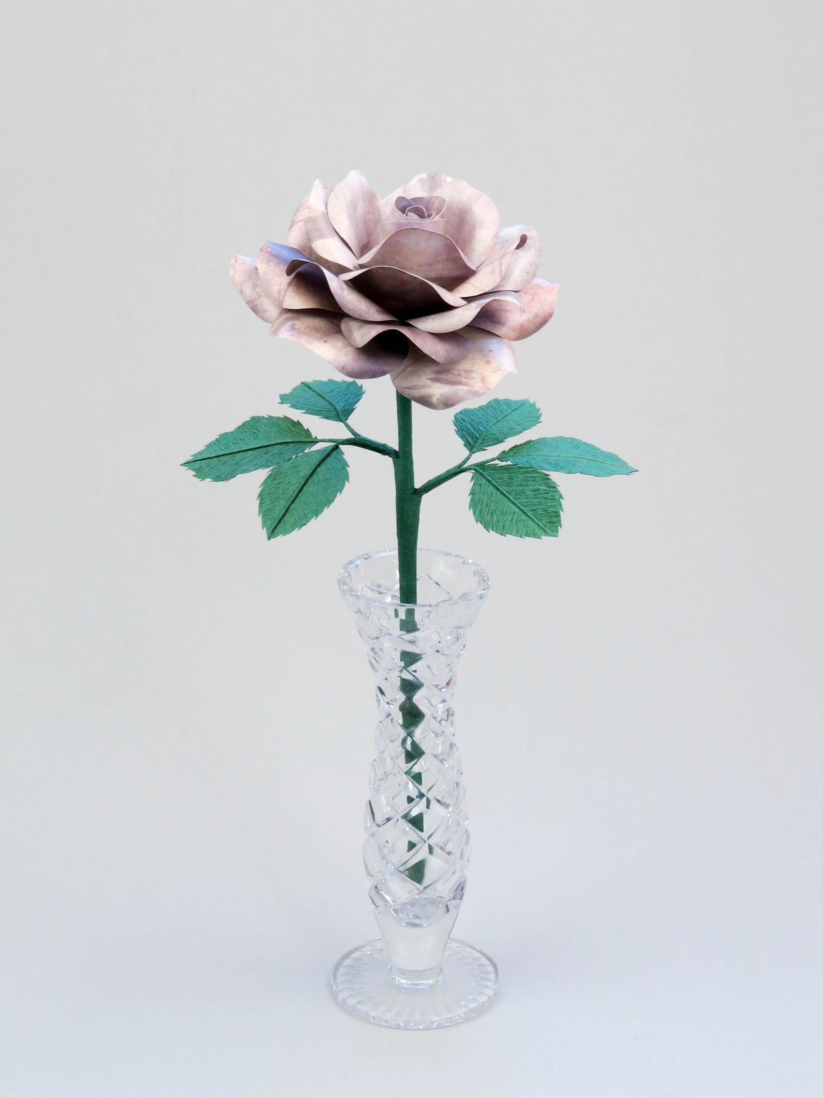 Light grey tin paper rose with six forest green leaves standing in a narrow glass vase set against a light grey background