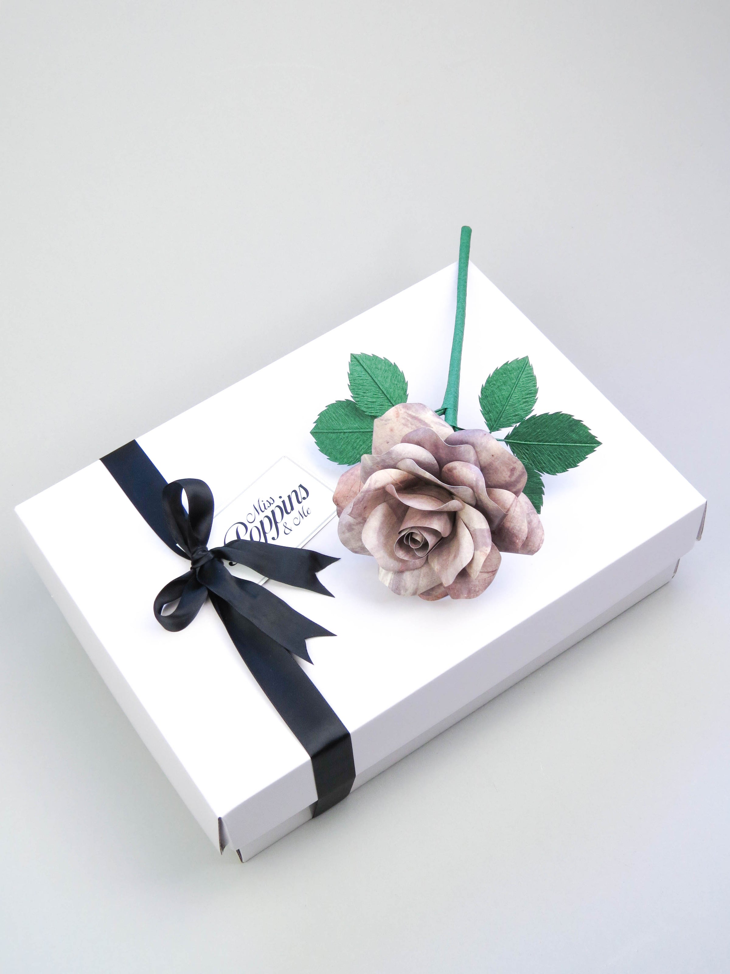 Grey tin paper rose lying diagonally on top of a luxury white gift box that has a black satin ribbon tied in a bow with a Miss Poppins and Me gift tag attached
