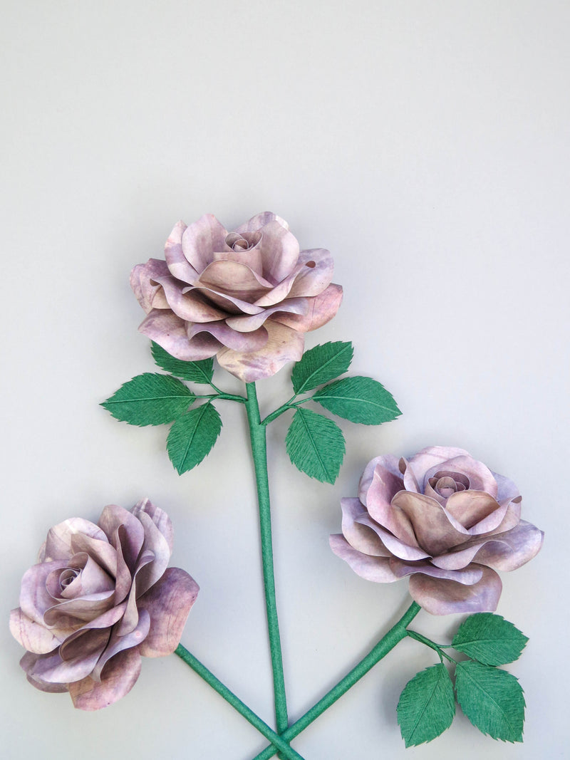 Three grey tin paper roses randomly lying next to each other on a light grey background. The left rose is leafless, the middle rose has six green leaves attached and the right rose has three leaves.