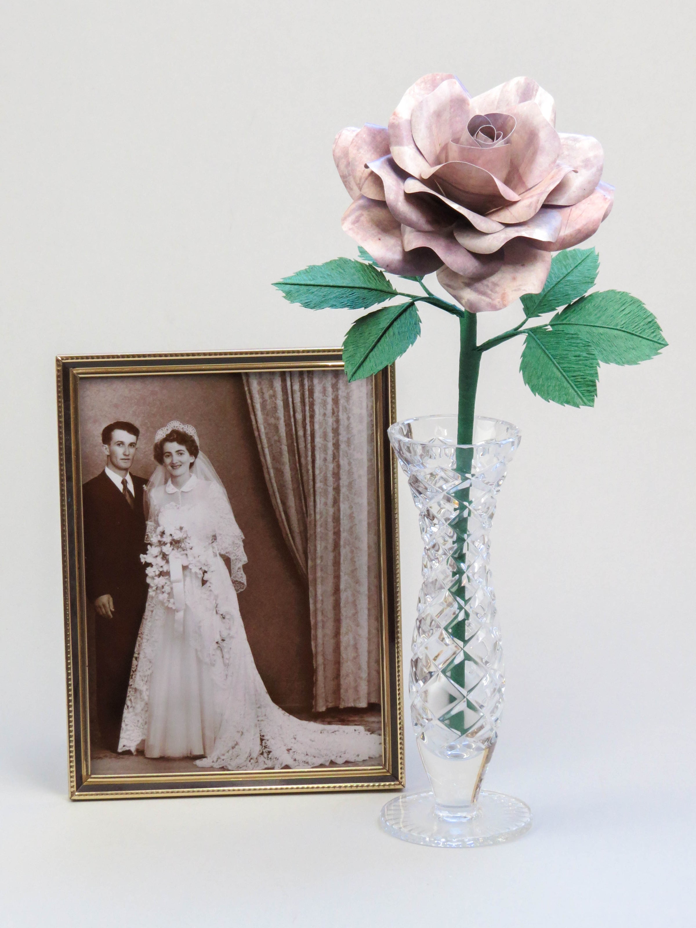 Grey tin paper rose with six forest green leaves standing in a slender glass vase with a thin gold framed sepia wedding photo of a happy vintage bride and groom standing beside it