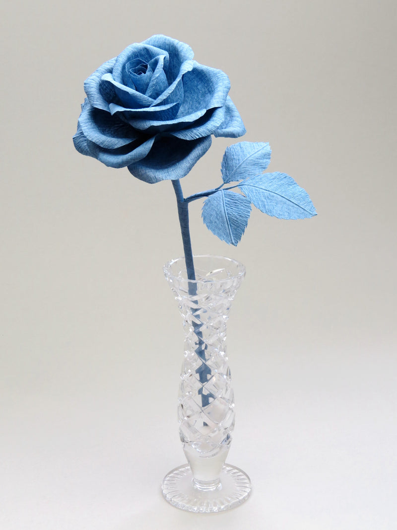 China blue crepe paper rose with three matching china blue leaves standing in a narrow glass vase against a light grey backdrop