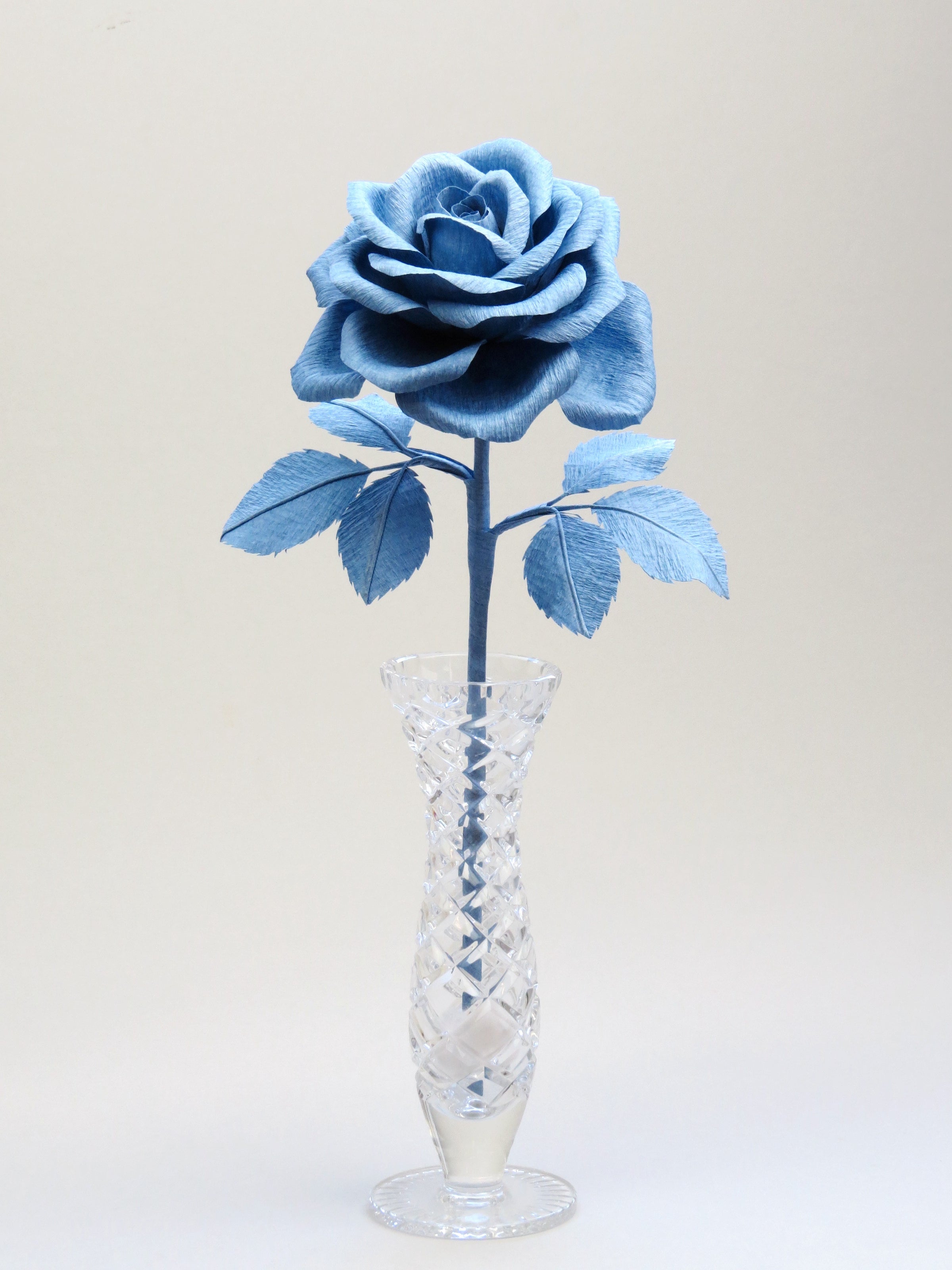 China blue crepe paper rose with six matching china blue leaves standing in a narrow glass vase set against a light grey background
