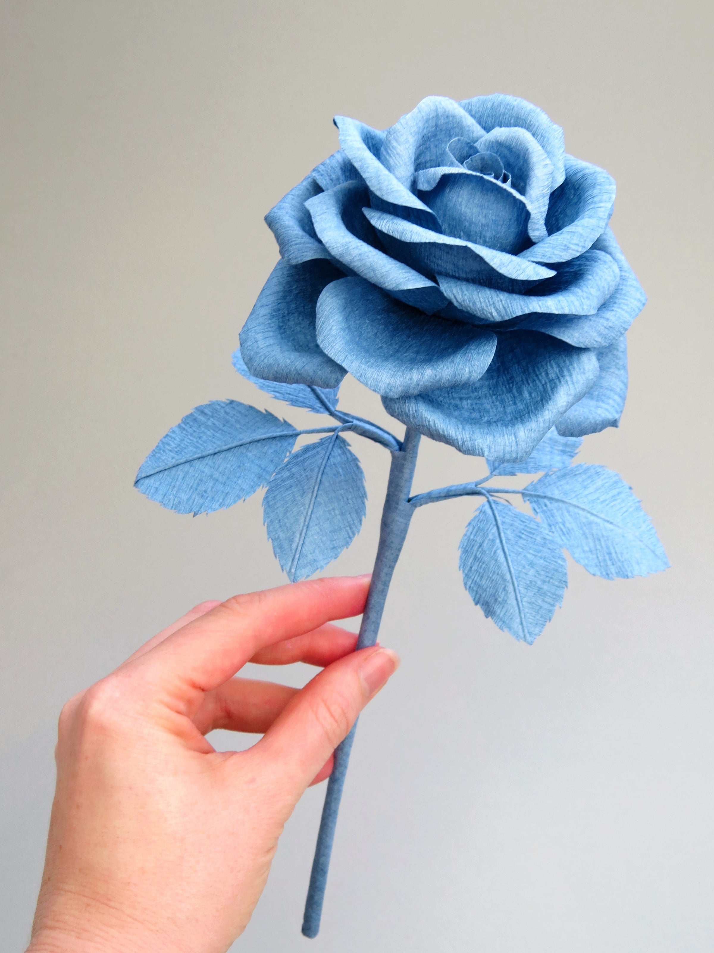 Pale white hand delicately holding the stem of a china blue paper rose with six matching china blue leaves