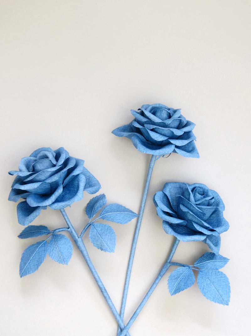 Three china blue crepe paper roses randomly lying next to each other on a light grey background. The left rose has six china blue leaves attached, the middle rose has no leaves and the right rose has three leaves.