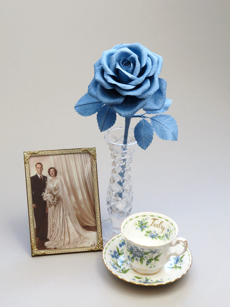 China blue crepe paper rose with six matching china blue leaves standing in a slender glass vase with a thin gold framed sepia wedding photo of a happy vintage bride and groom standing to the left, with a vintage floral tea cup and saucer placed to the right