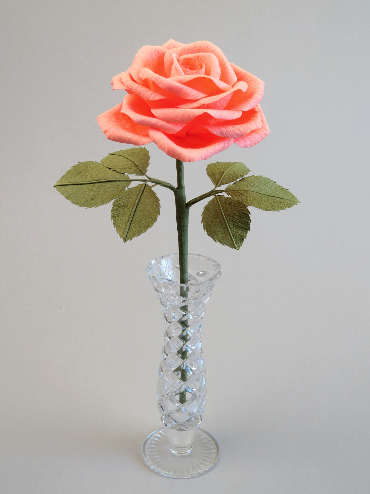 Coral pink crepe paper rose with six olive green leaves standing in a narrow glass vase set against a light grey background