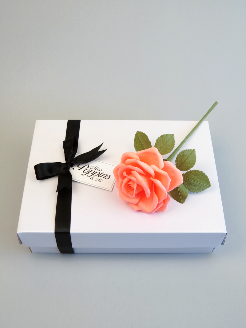Coral pink crepe paper rose lying diagonally on top of a luxury white gift box that has a black satin ribbon tied in a bow with a Miss Poppins and Me gift tag attached