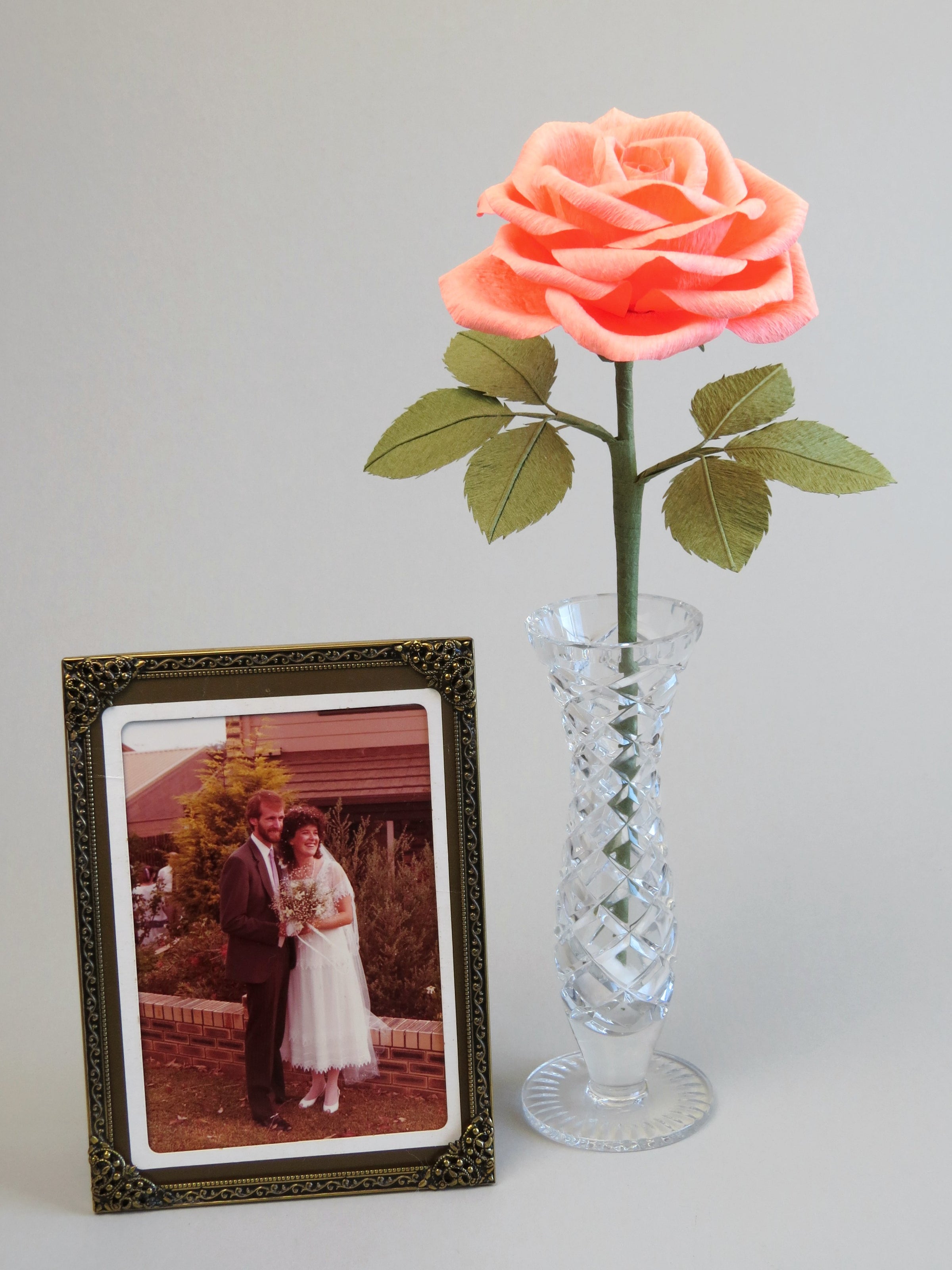Coral pink crepe paper rose with six olive green leaves standing in a slender glass vase with a thin gold framed retro wedding photo of a happy 1980s does 1920s inspired bride and groom in a garden standing beside it
