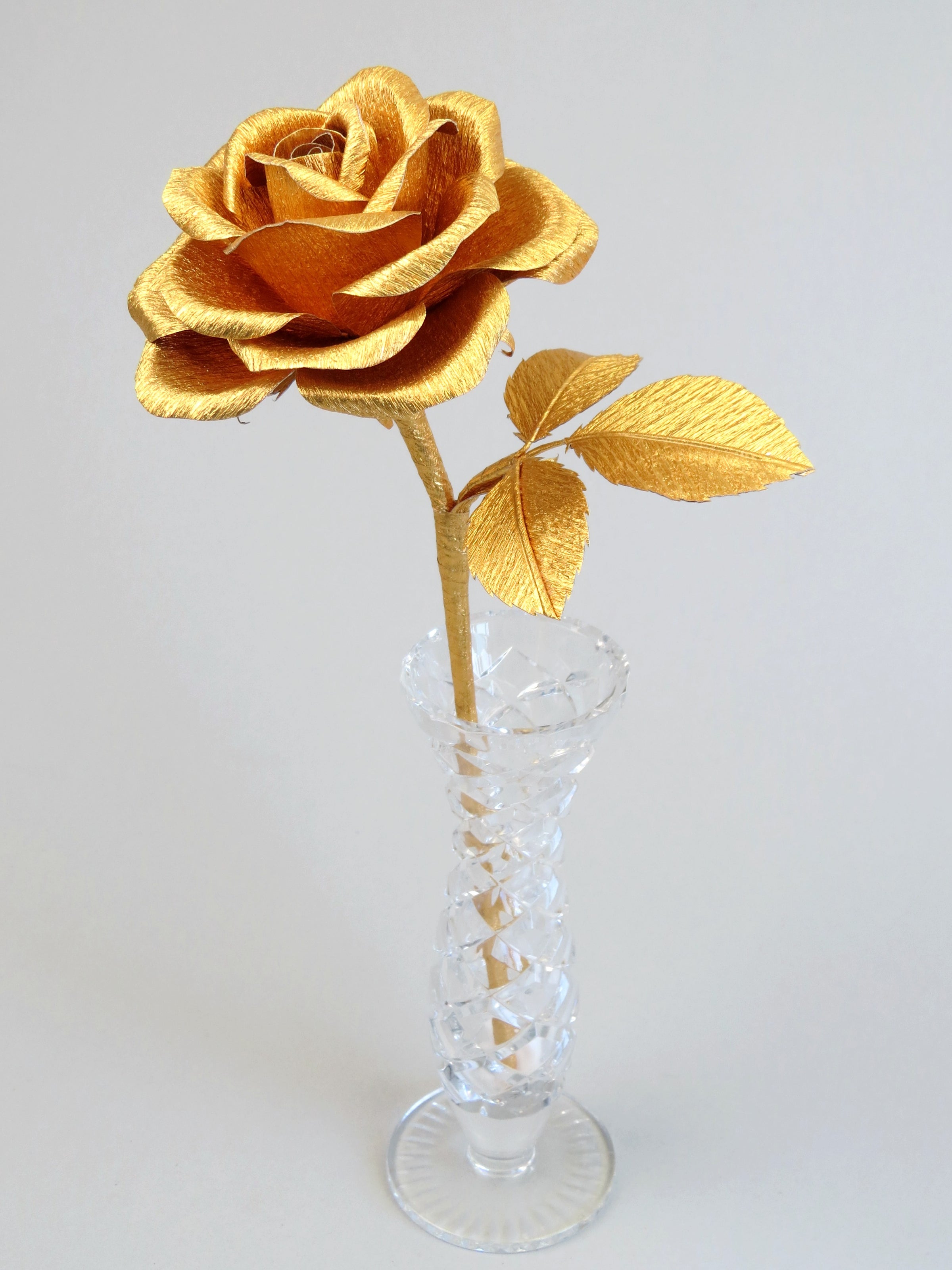 Gold crepe paper rose with three gold leaves standing in a narrow glass vase against a light grey backdrop