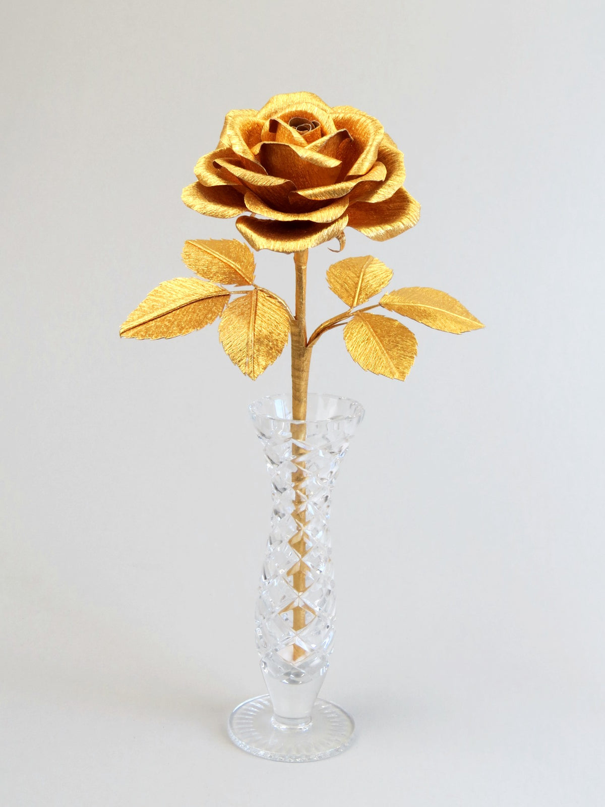 Gold crepe paper rose with six gold leaves standing in a narrow glass vase set against a light grey background