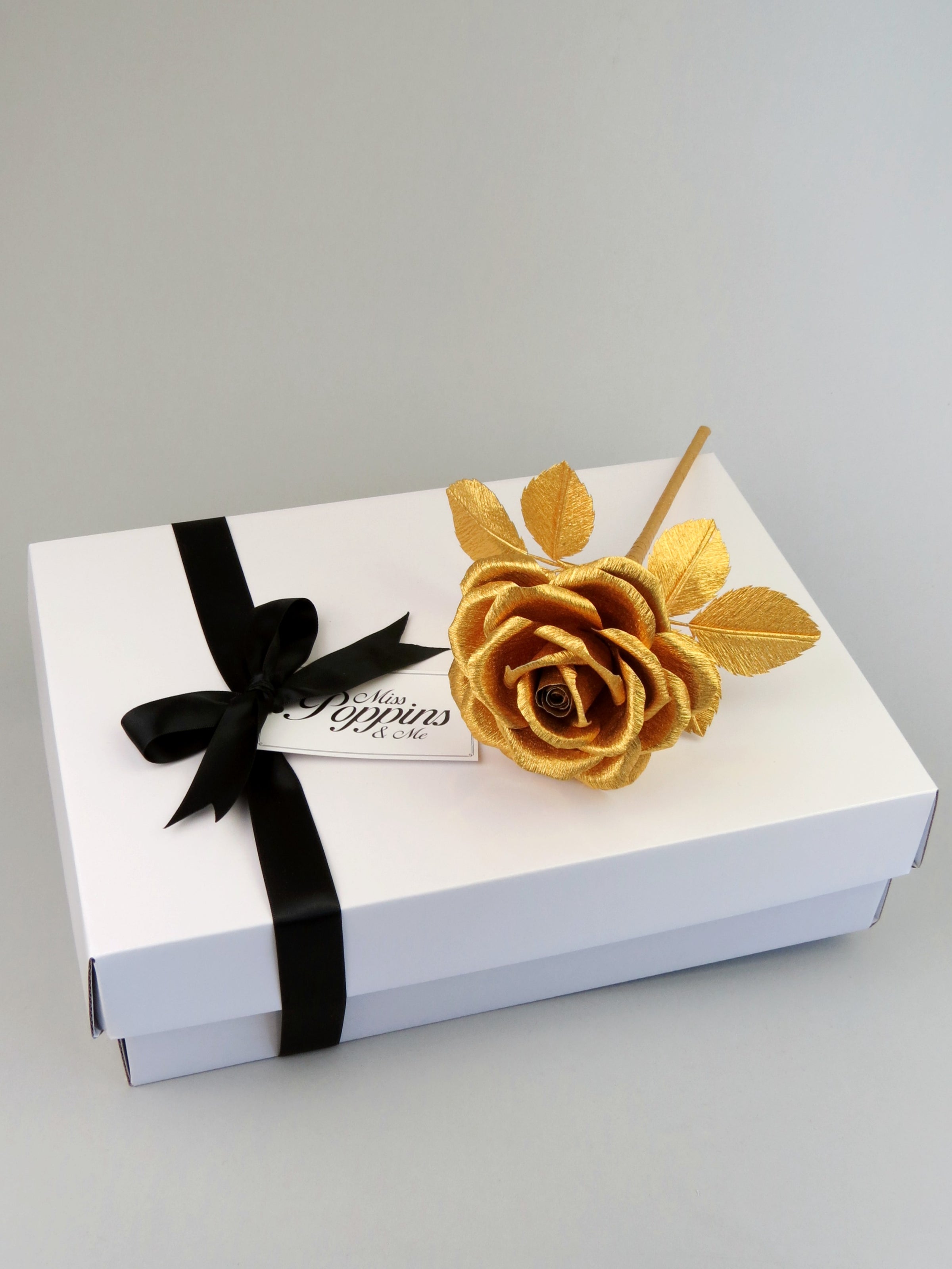 Gold crepe paper rose lying diagonally on top of a luxury white gift box that has a black satin ribbon tied in a bow with a Miss Poppins and Me gift tag attached