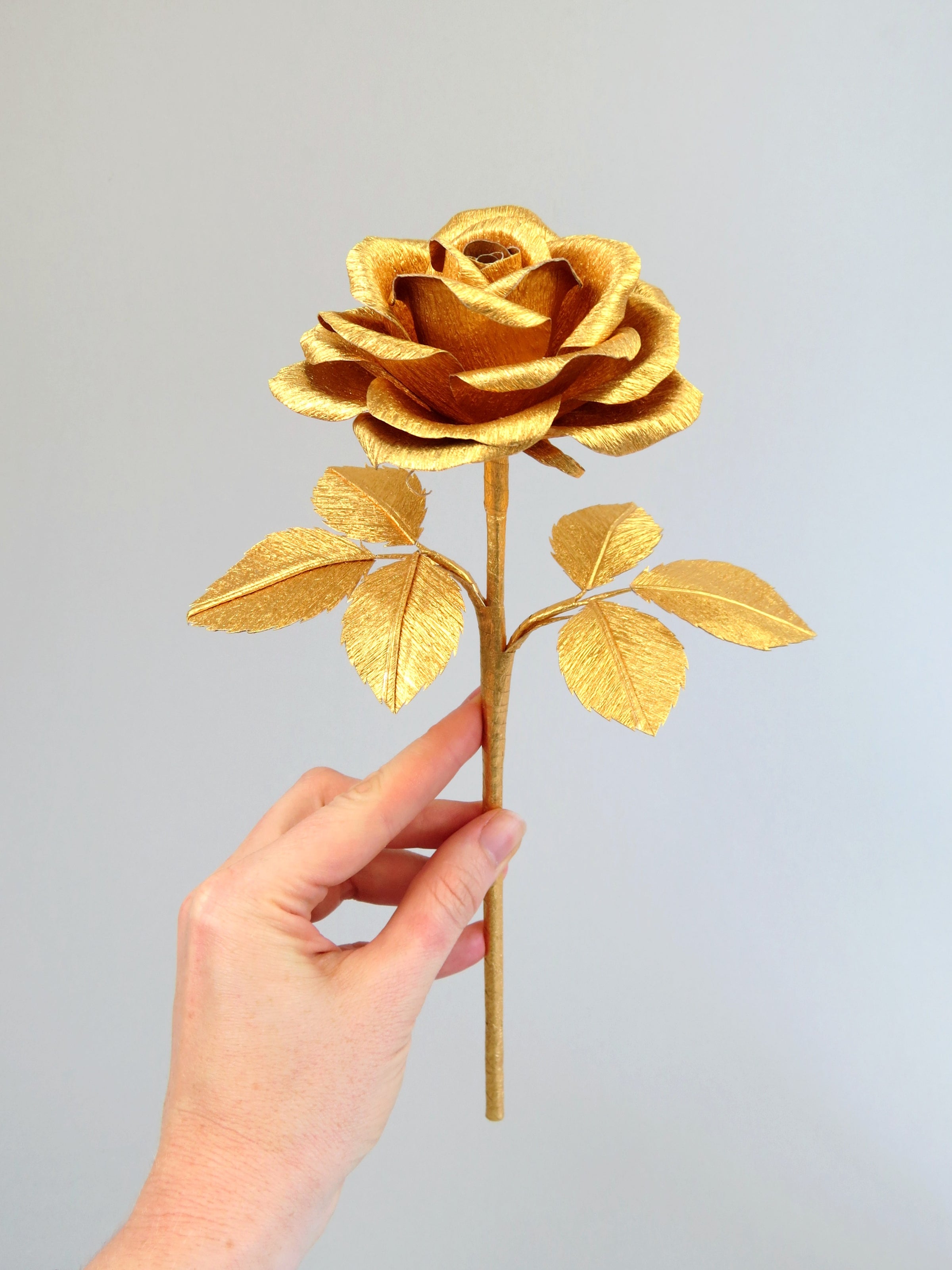 Pale white hand delicately holding the stem of a gold paper rose with six gold leaves
