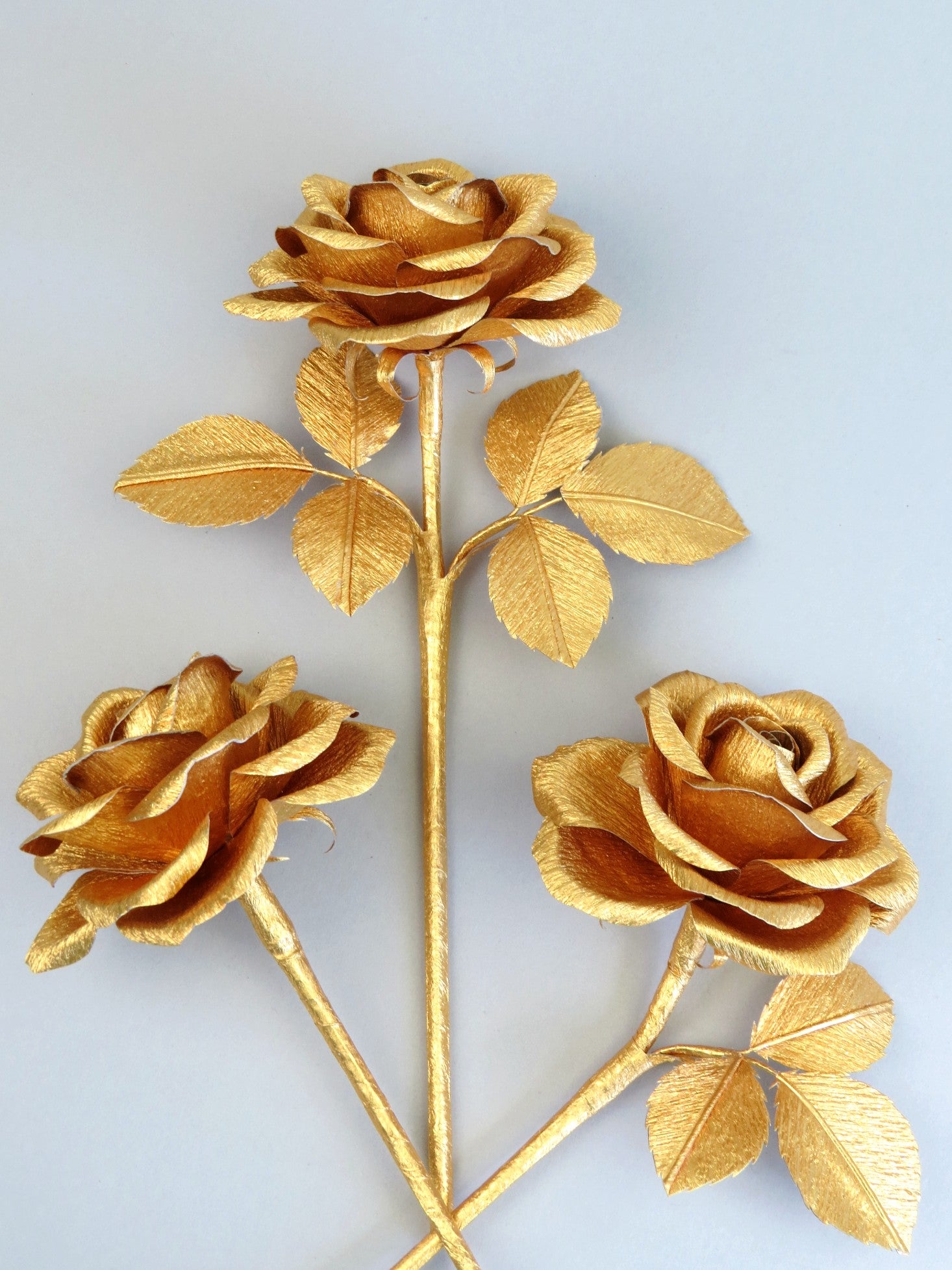 Three gold crepe paper roses randomly lying next to each other on a light grey background. The left rose is leafless, the middle rose has six gold leaves attached and the right rose has three gold leaves.