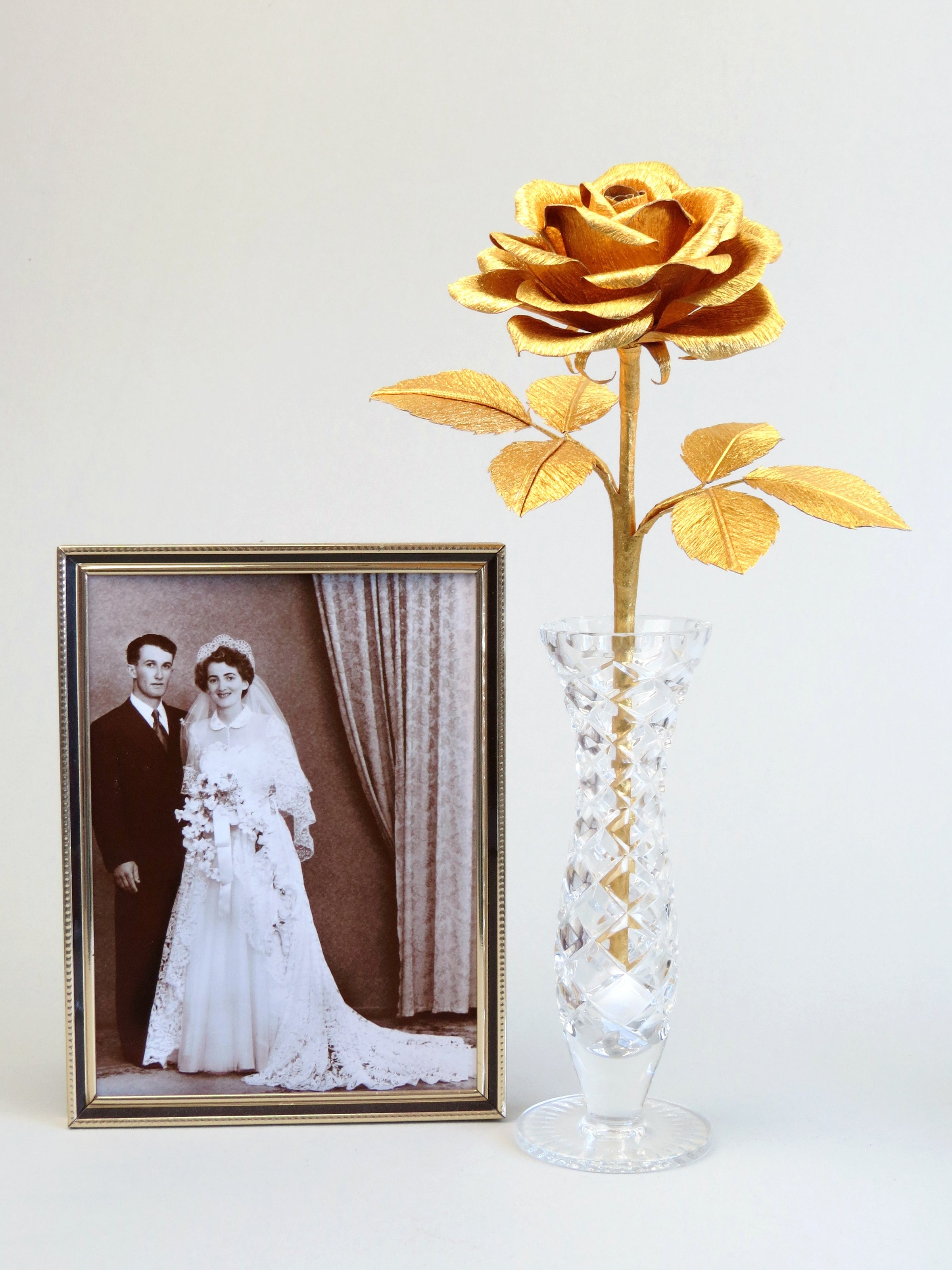 Gold crepe paper rose with six gold leaves standing in a slender glass vase with a thin gold framed sepia wedding photo of a happy vintage bride and groom standing to the left of the vase