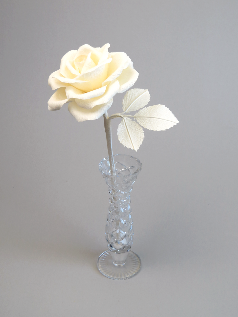 Ivory crepe paper rose with three ivory leaves standing in a narrow glass vase against a light grey backdrop