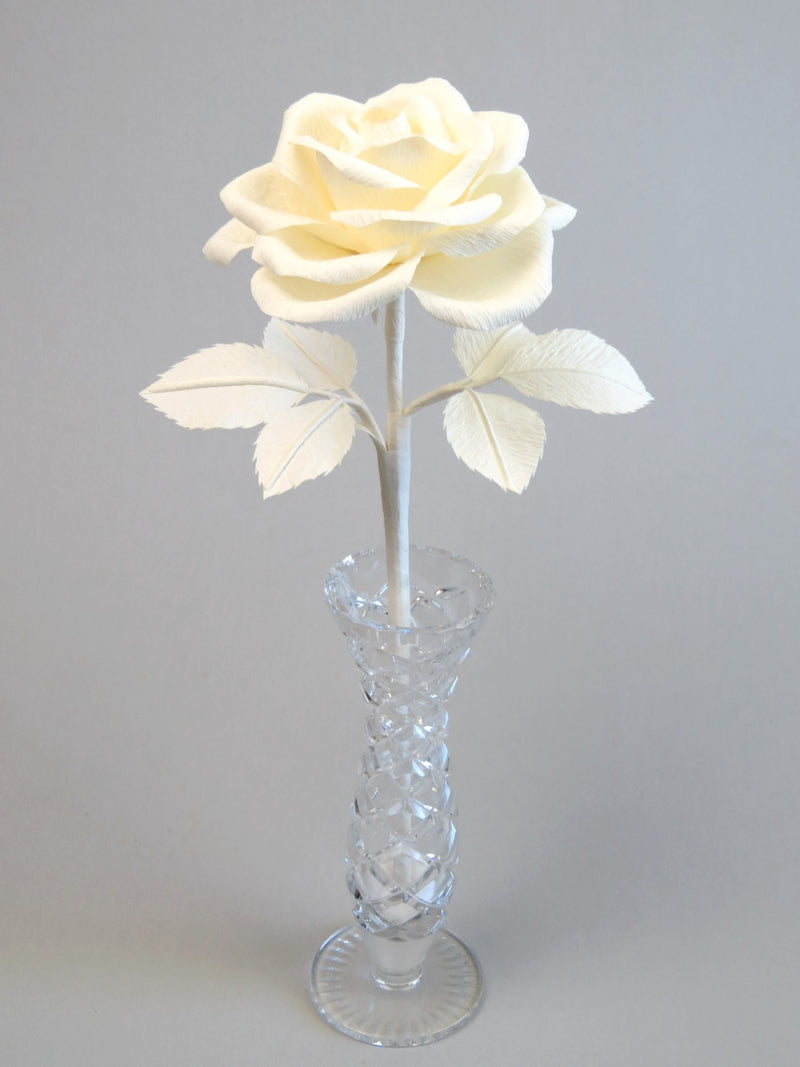 Ivory crepe paper rose with six ivory leaves standing in a narrow glass vase set against a light grey background