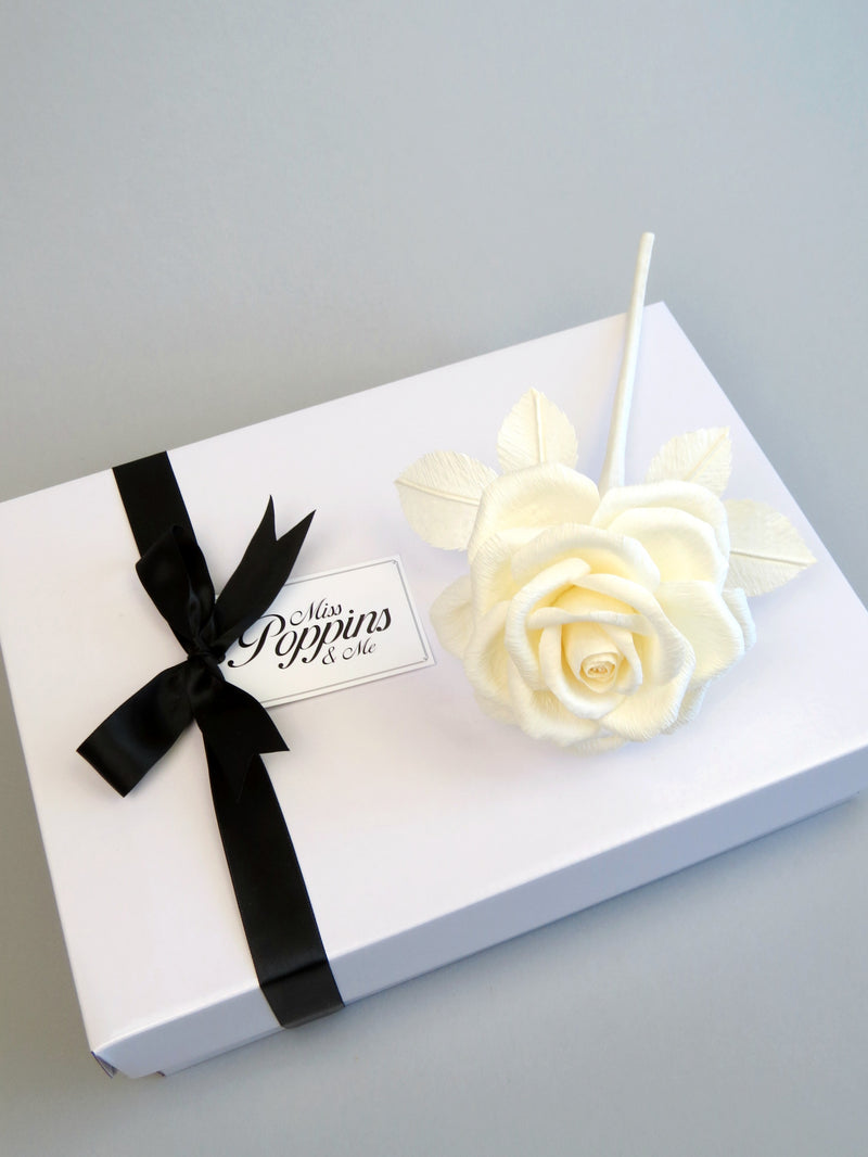 Ivory crepe paper rose lying diagonally on top of a luxury white gift box that has a black satin ribbon tied in a bow with a Miss Poppins and Me gift tag attached