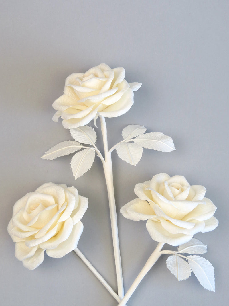 Three ivory crepe paper roses randomly lying next to each other on a light grey background. The left rose is leafless, the middle rose has six ivory leaves attached and the right rose has three leaves.
