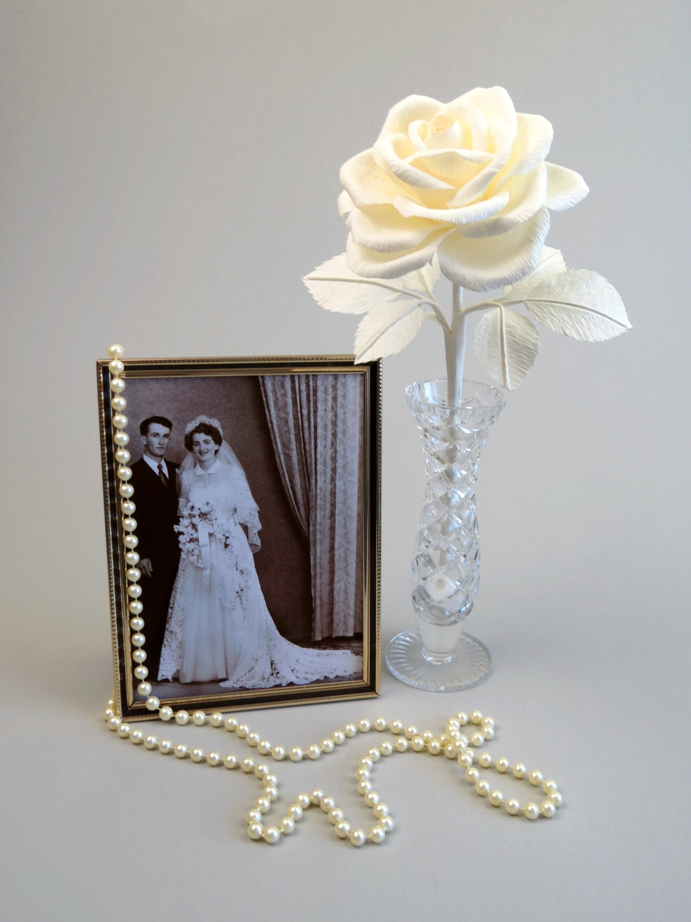 Ivory crepe paper rose with six ivory leaves standing in a slender glass vase with a thin gold framed sepia wedding photo of a happy vintage bride and groom standing beside it with a necklace of pearls draped over the frame