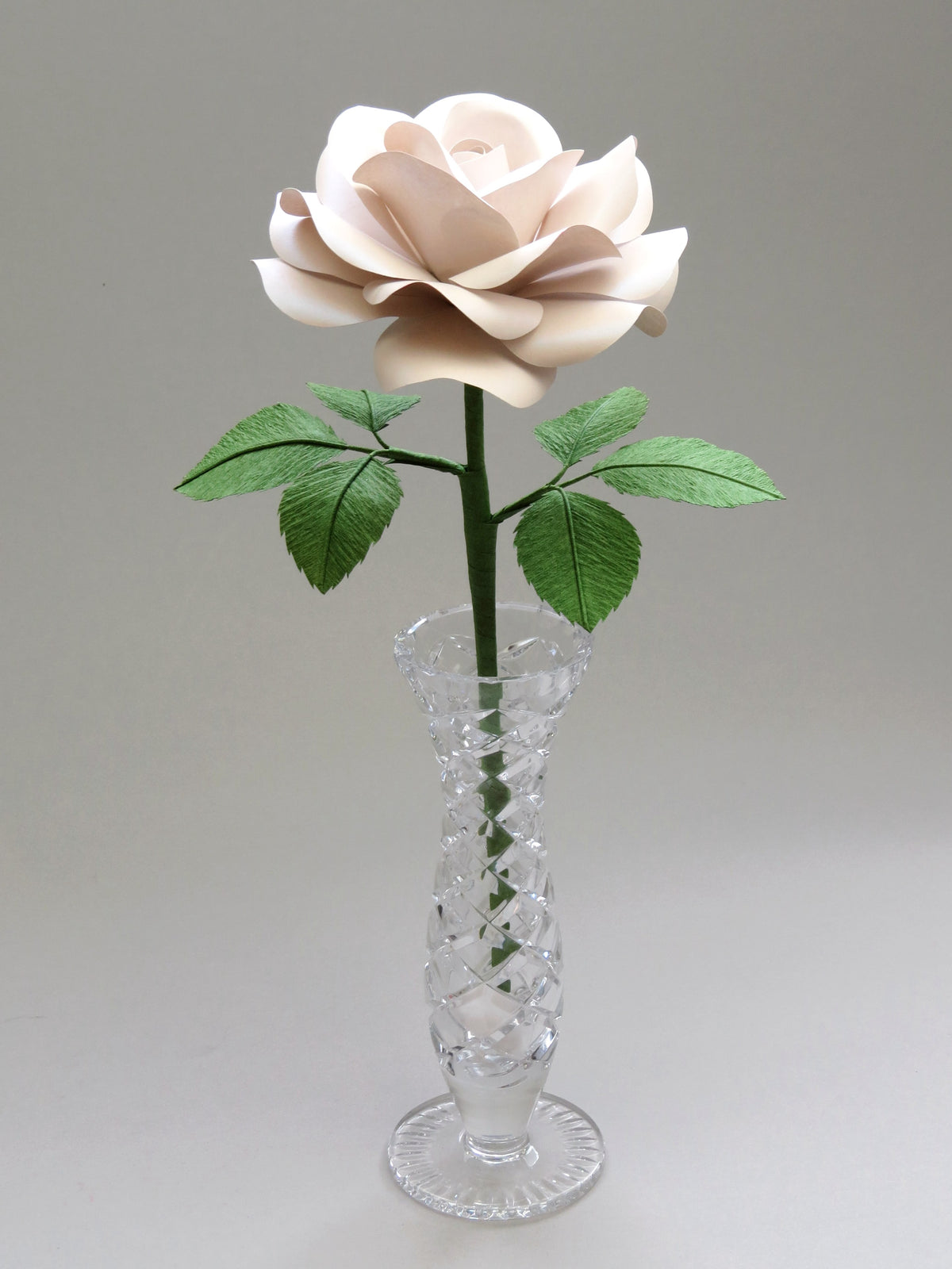 Pearl paper rose with six ivy green crepe paper leaves standing in a narrow glass vase set against a light grey background